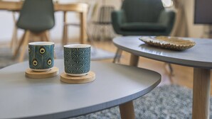 Una mesa redonda con dos tazas decorativas sobre posavasos, una pequeña bandeja con un adorno y un sillón verde al fondo.
