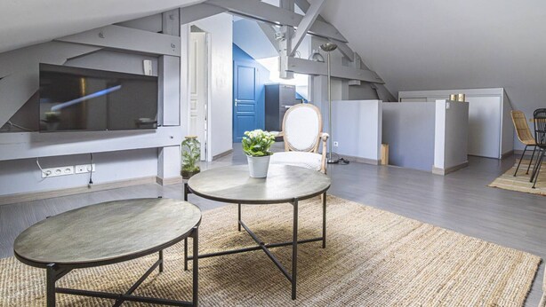 Um apartamento loft moderno com teto alto, vigas aparentes, um amplo claraboia, área de jantar com mesa e cadeiras redondas, televisão na parede e uma porta azul ao fundo.