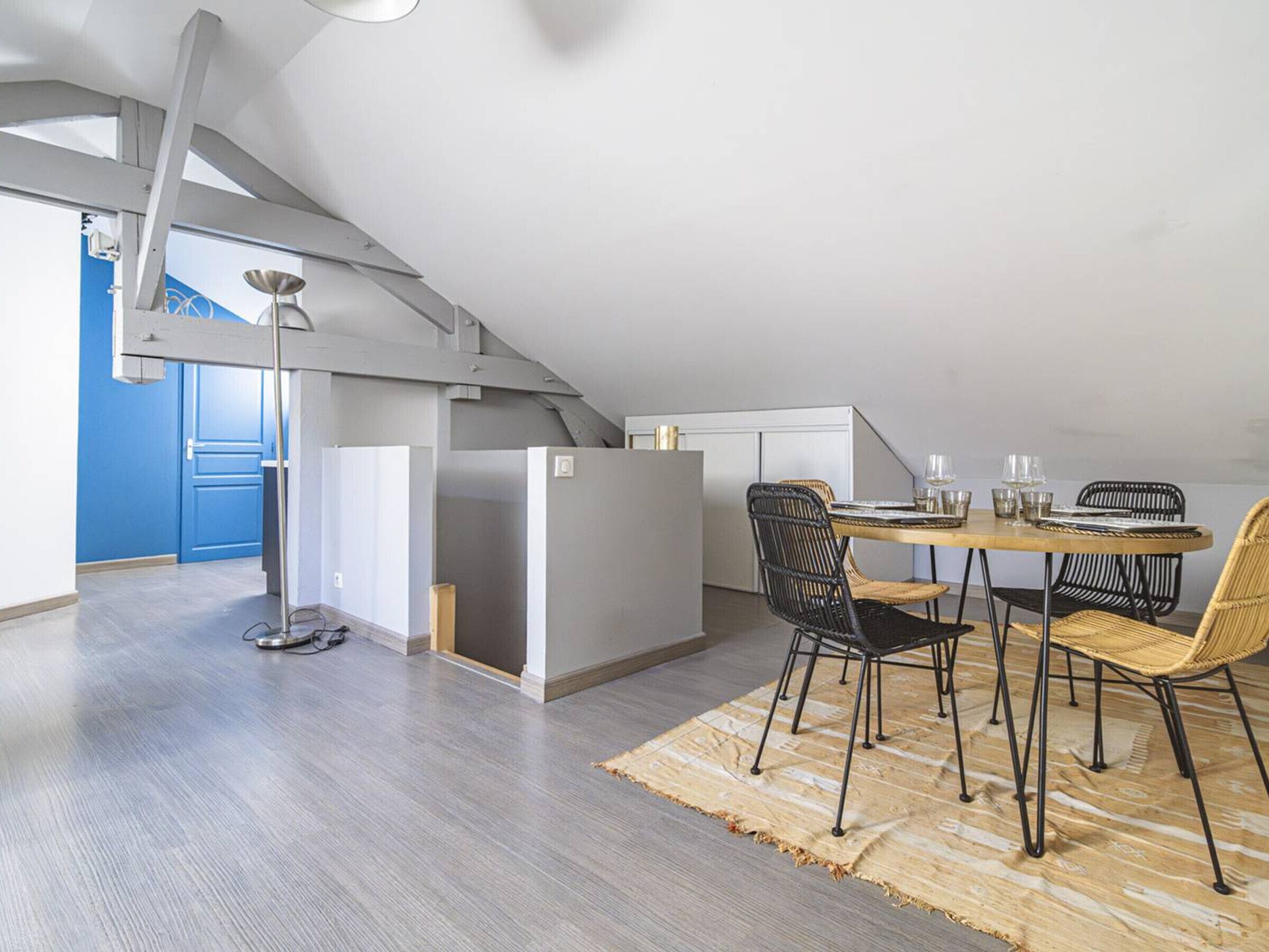 A modern loft apartment with a dining area featuring a round table and black chairs, a visible kitchen area, and a high ceiling with exposed beams.