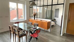 Un espace repas moderne avec une table en verre, des chaises en bois et un fauteuil rouge. Un buffet en bois, doté de tiroirs, est adossé au mur, et une grande fenêtre offre une vue sur l’extérieur.
