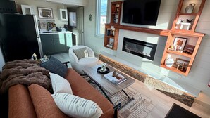 A modern living room with a sofa, coffee table, and fireplace. There is a kitchen area visible in the background. - Green Drake Tiny Home (Pray)