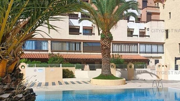 A swimming pool with a palm tree and a building in the background. - Charming apartment with balcony, pool view - 5 guests (Gruissan)