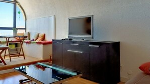 A compact living space with a kitchenette, a glass-top table, a sofa, and a television mounted on the wall. - Charming apartment with balcony, pool view - 5 guests (Gruissan)