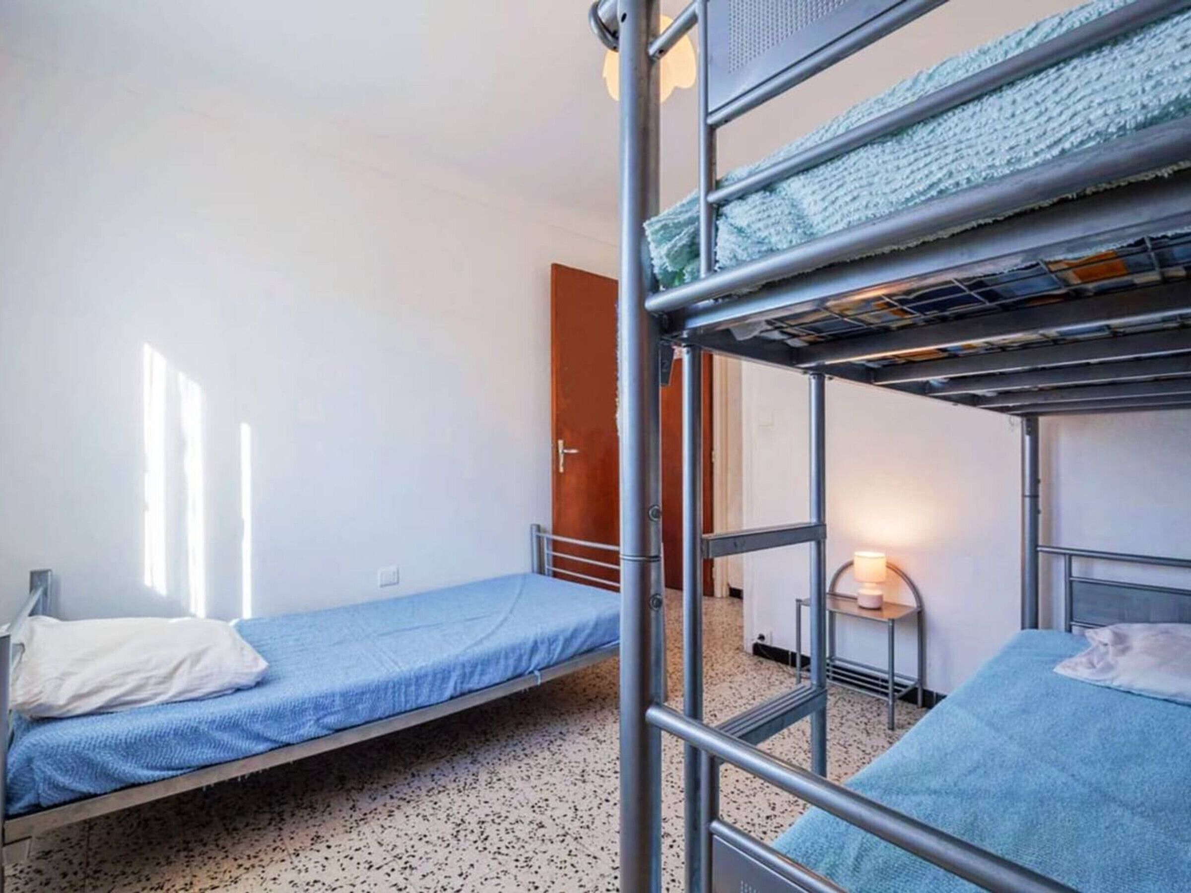 A dormitory room with bunk beds, a small bedside table with a lamp, and a door leading to another room.