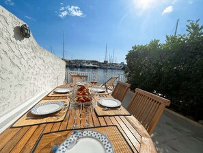 Mesa de madera preparada para comer con platos, vasos y un bol de fruta, con vista a un puerto deportivo con barcos.