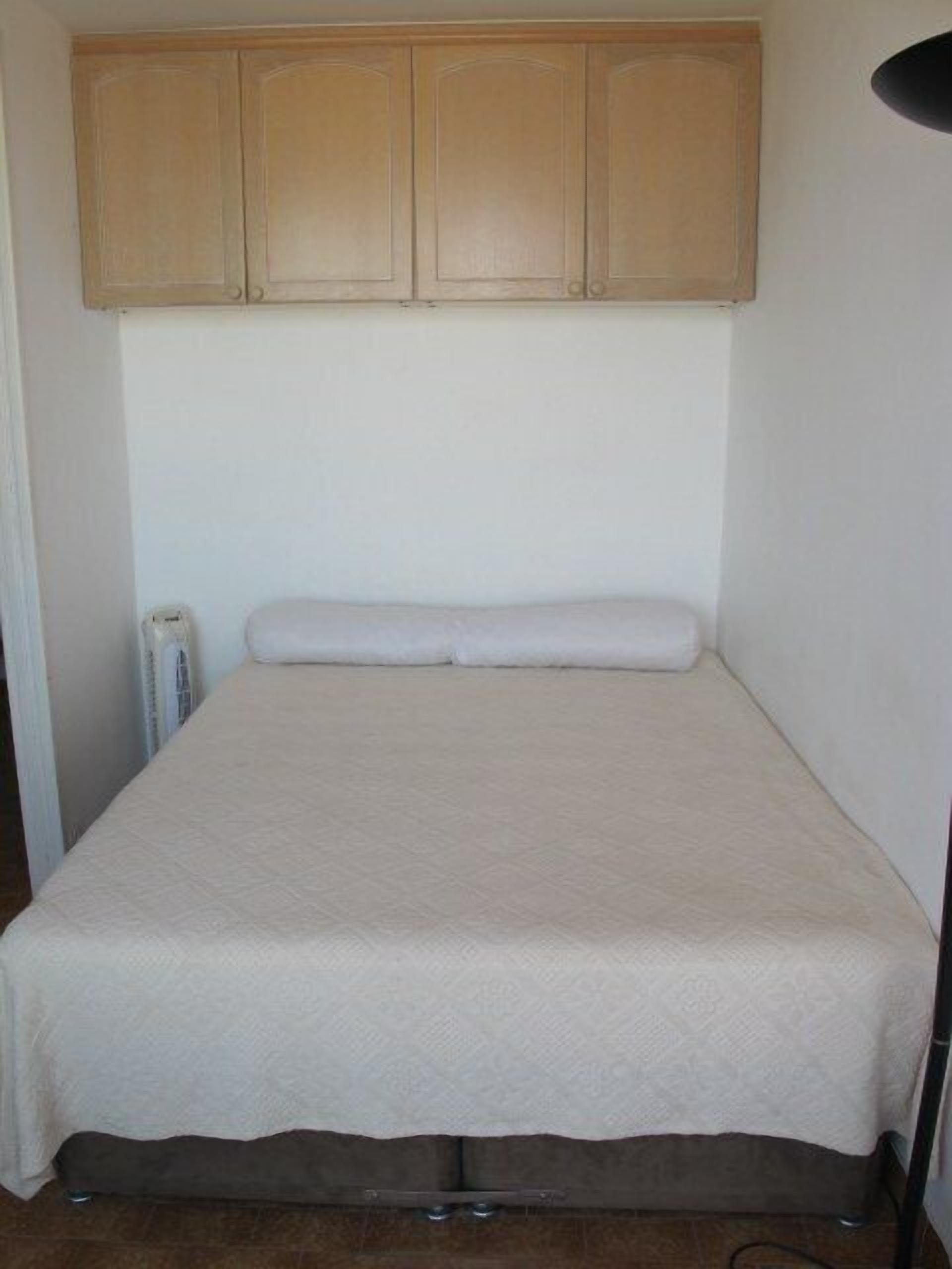 Une petite chambre avec un lit simple, recouverte d’une housse de couette blanche unie, avec des placards en bois au-dessus et un radiateur sur le côté gauche.