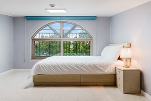 Ein Schlafzimmer mit einem großen Bogenfenster, einem Bett mit weißer Bettwäsche, einem Nachttisch mit einer Lampe und einem Fischgrätenparkettboden.