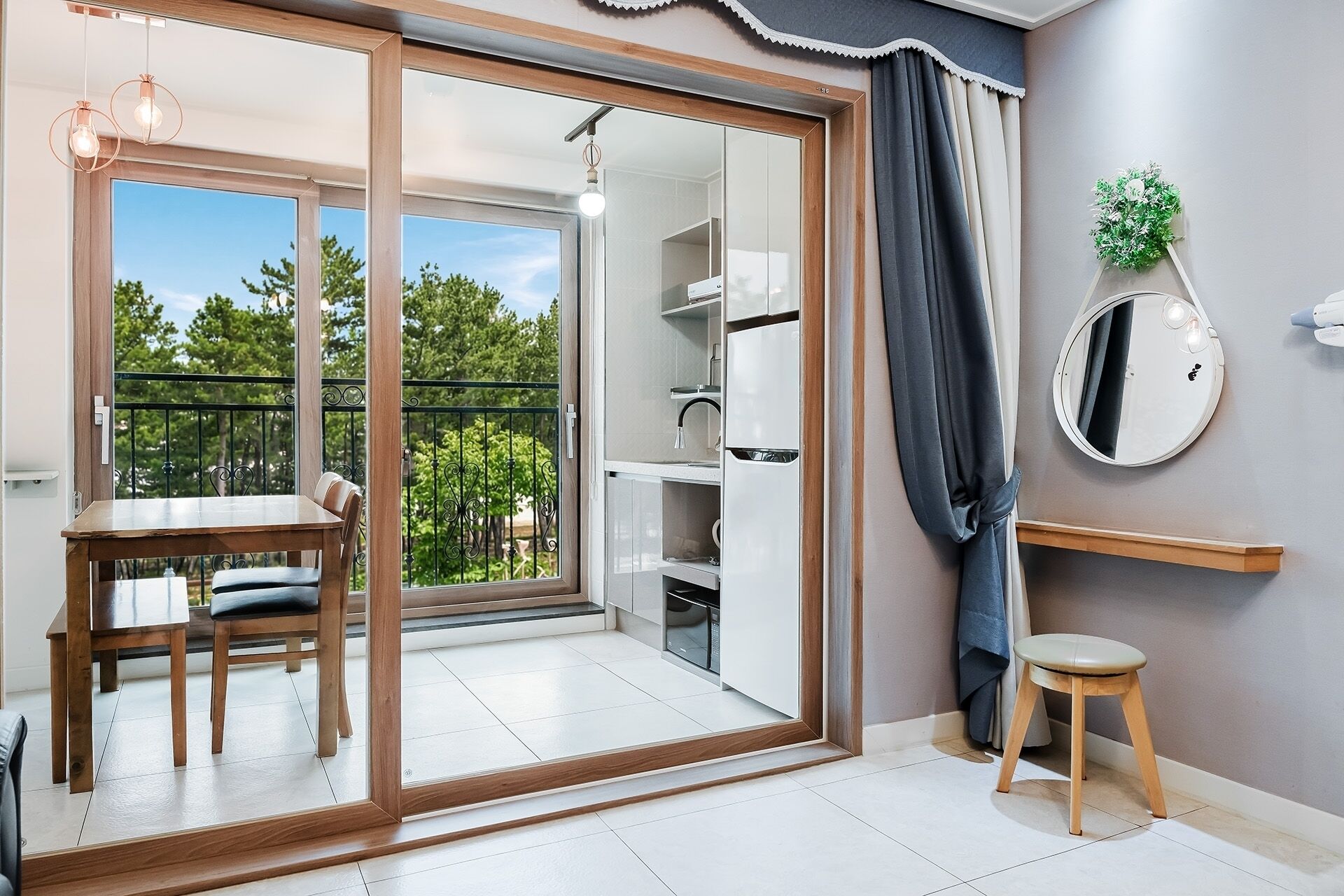 Una habitación con una puerta corrediza de vidrio que da a un balcón, una mesa de madera con sillas, un taburete pequeño, un espejo redondo y una estantería con una planta.
