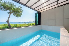 A modern indoor swimming pool with a clear view of the sea and a tree outside.