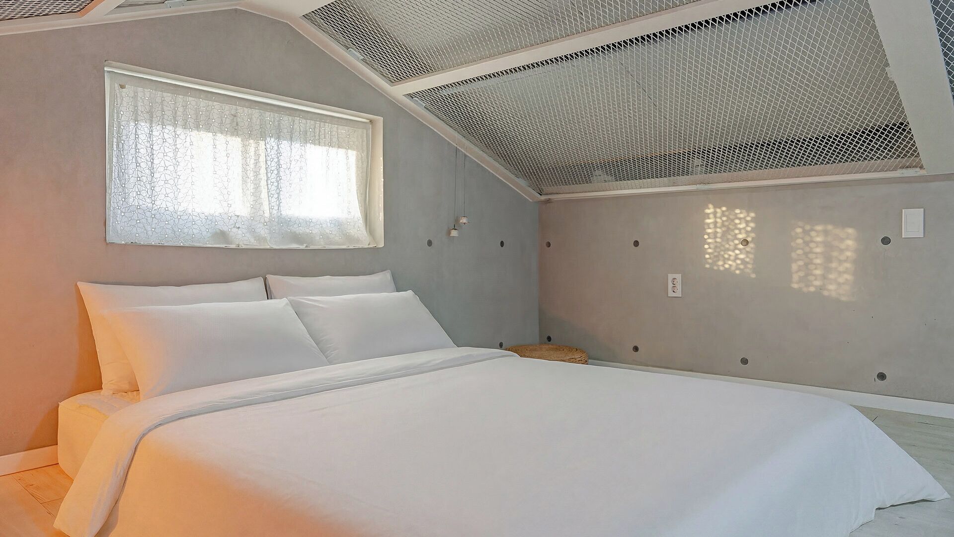 Un dormitorio con cama, una ventana con cortinas traslúcidas y un techo inclinado con diseño de malla.