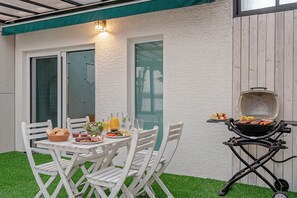 Un patio exterior con una mesa blanca lista para una comida, una parrilla y un toldo verde.