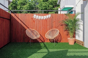 Un espacio al aire libre cercado, con césped artificial, dos sillas de mimbre y una planta en maceta.