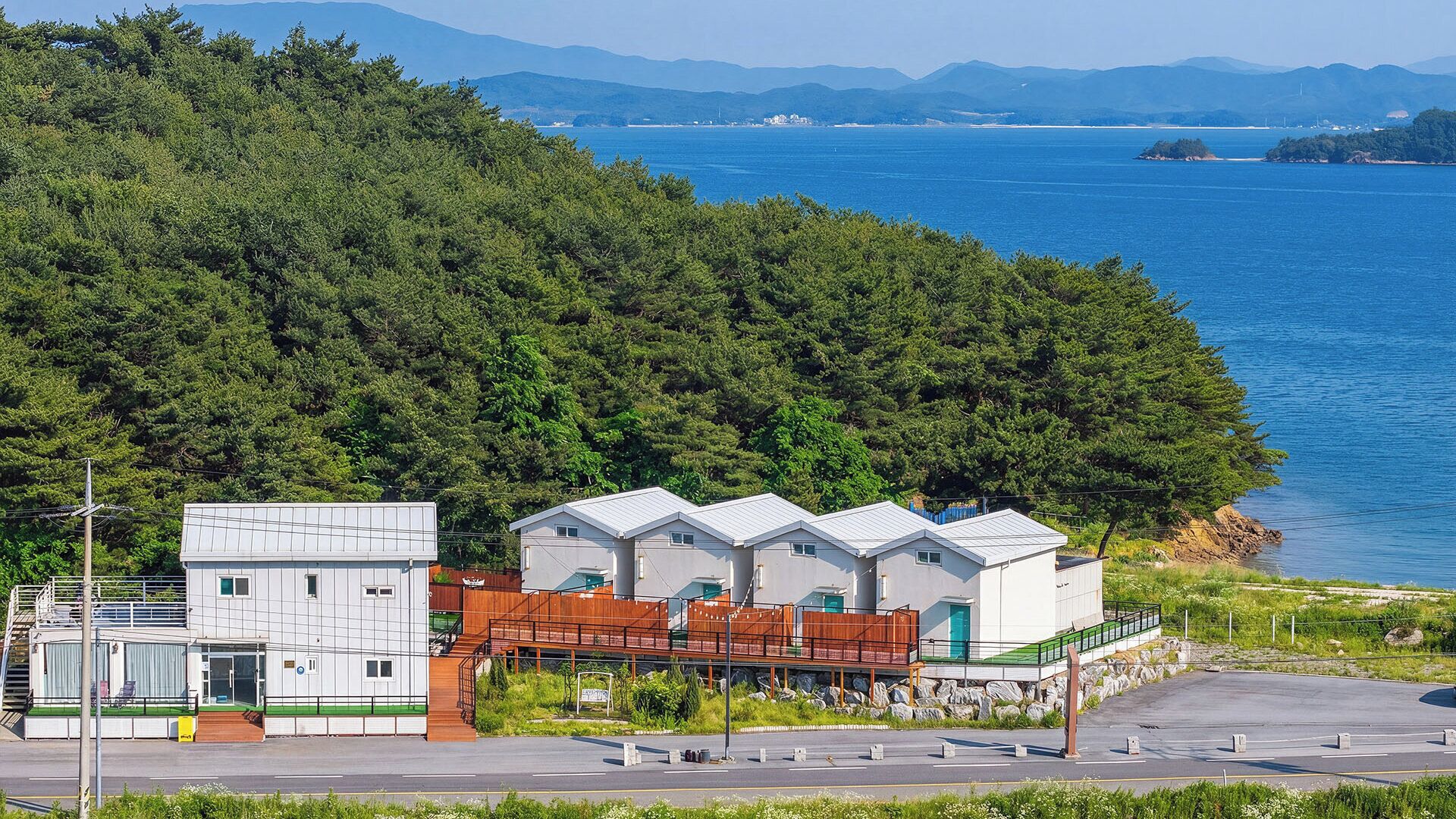A coastal building with a parking area, surrounded by dense forest and a view of the sea.