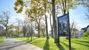 A park with a paved walkway, trees with autumn leaves, and a poster on a post. - Bel appartement vue mer (Camiers)