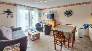 A living room with a sofa, armchair, and dining table with chairs. There is a television on a stand, a clock on the wall, and a window with curtains. - Bel appartement vue mer (Camiers)