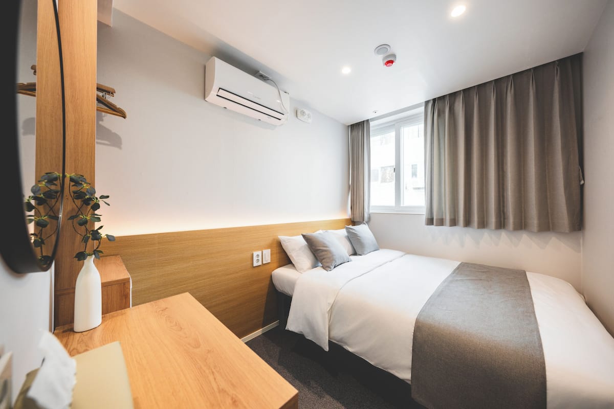 Una habitación de hotel con una cama, cabecera de madera, mesita de noche, un jarrón blanco con una planta y una ventana con cortinas.