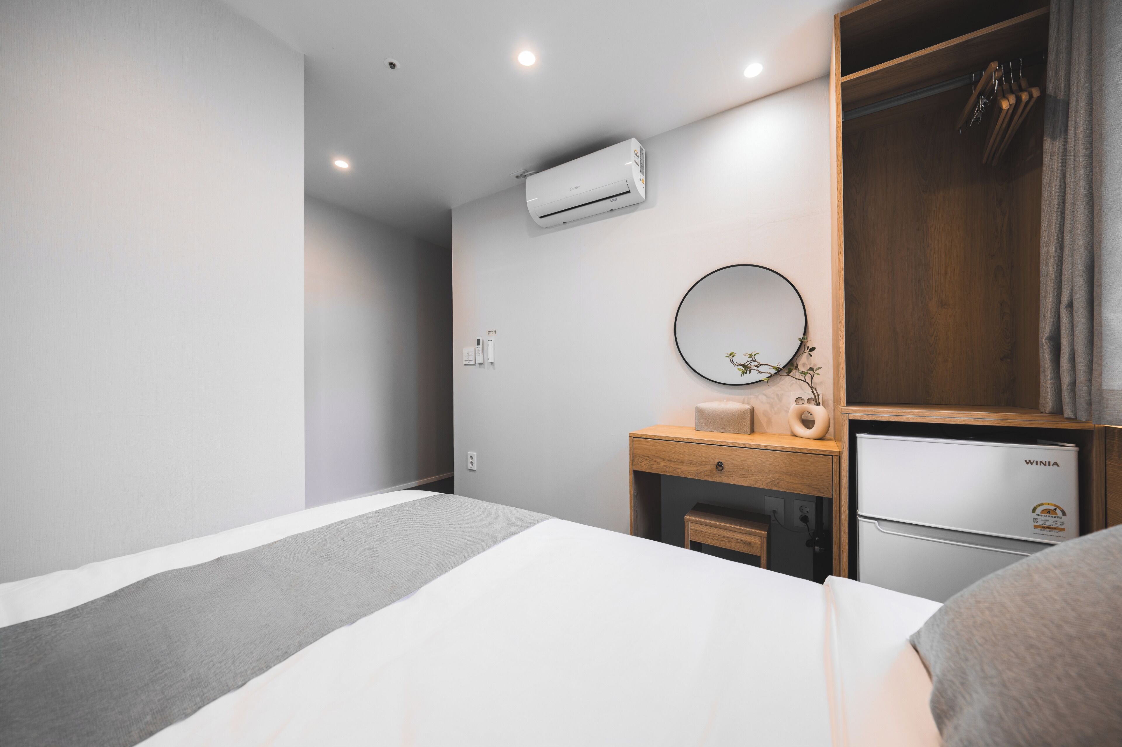 A modern hotel room with a bed, a wooden wardrobe, a small desk with a mirror, and a built-in mini-fridge.