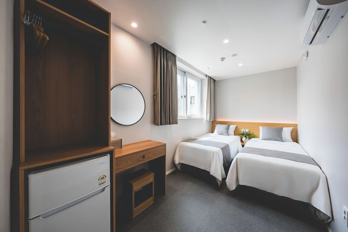 Habitación de hotel con dos camas, un escritorio, un refrigerador pequeño, un espejo y una ventana con cortinas.