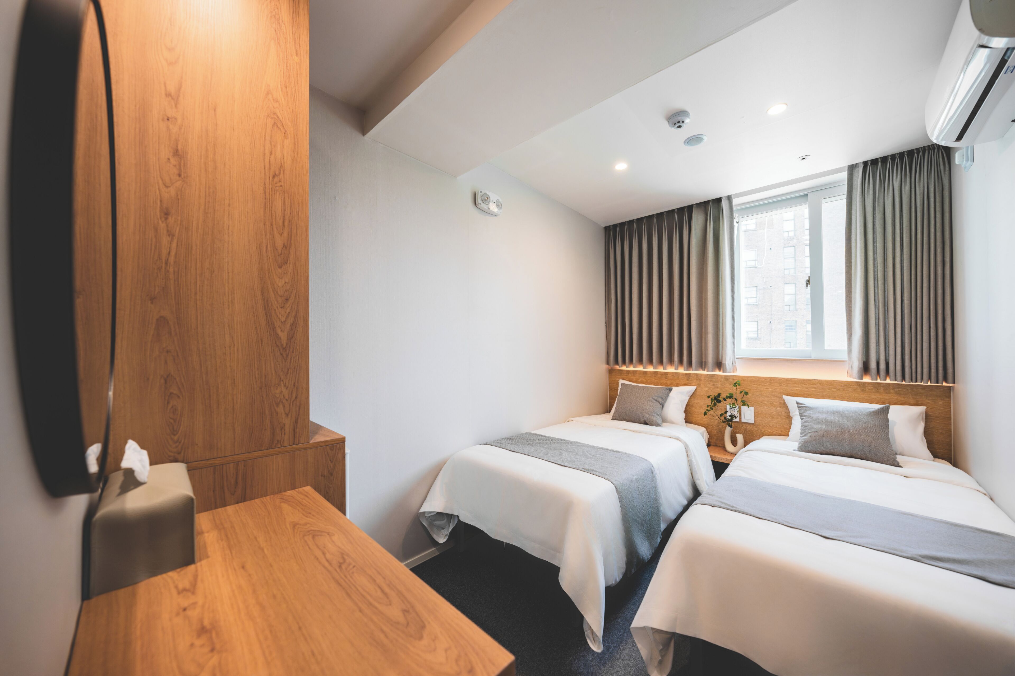 A hotel room with two beds, a wooden desk, a mirror, and a window with curtains.