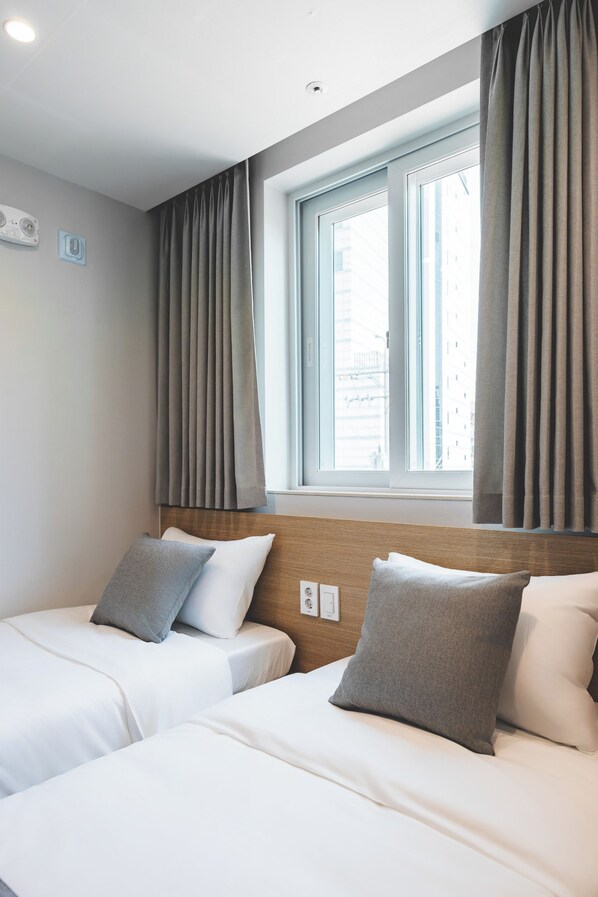 Hotellihuone, jossa on kaksi sänkyä, ikkuna, jossa on verhot, puinen sängynpääty ja näkyvissä oleva pistorasia seinässä.
