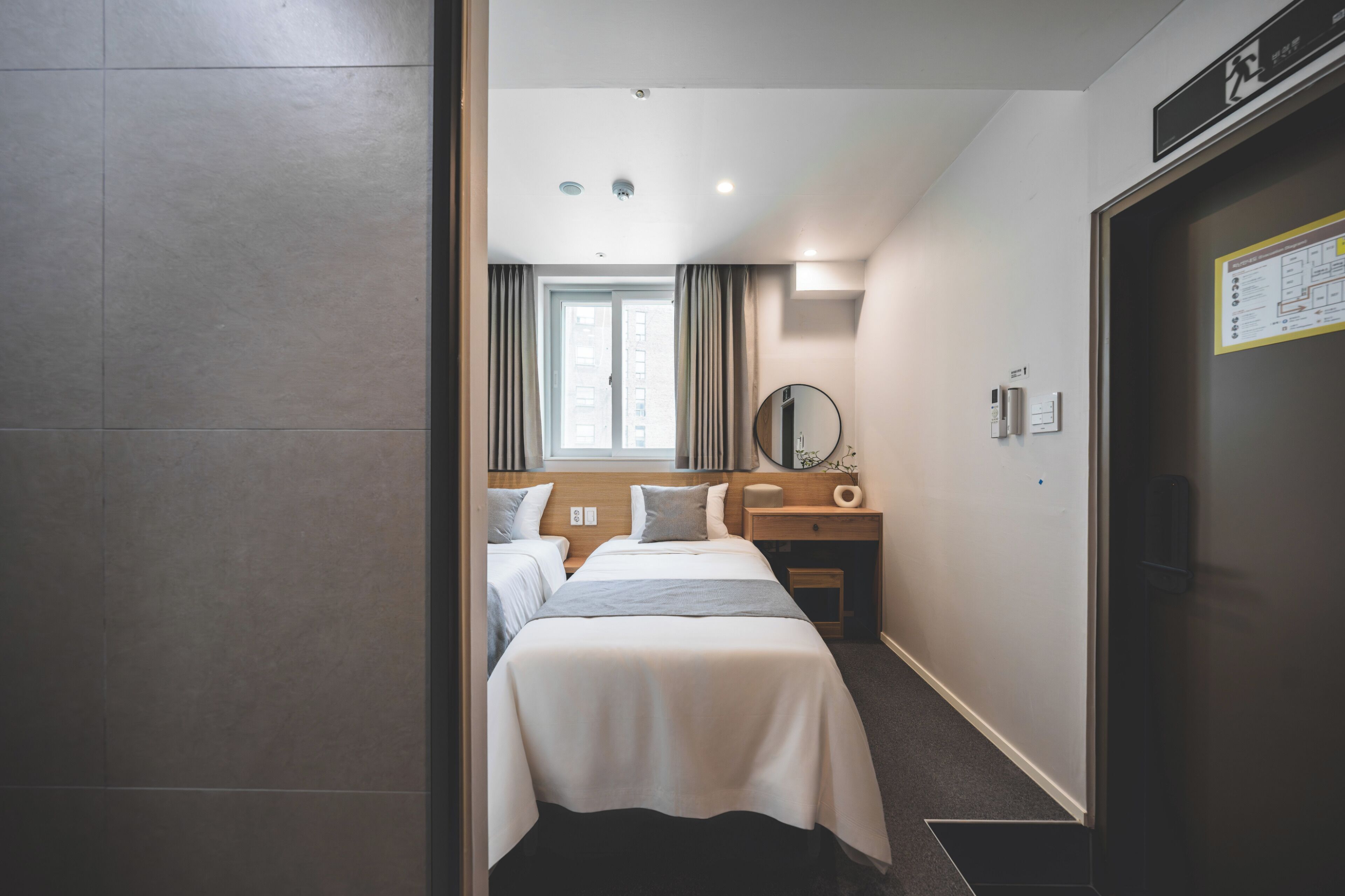 A hotel room with two beds, a bedside table, a mirror, a window with curtains, and a door with a sign.