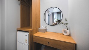 Ein kleines Zimmer mit Schreibtisch aus Holz, einem Spiegel, einem kleinen, weißen Kühlschrank und einer Vase mit Pflanze.