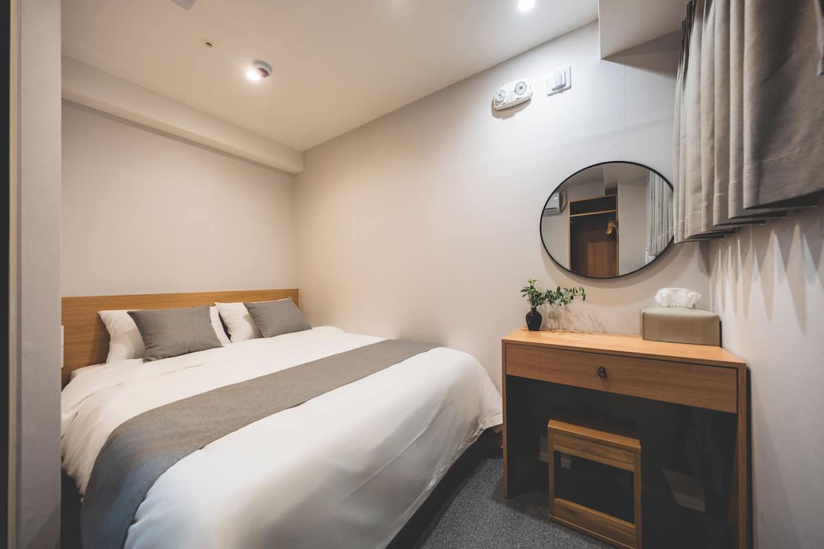 Una habitación de hotel con cama, mesita de noche con espejo, una planta pequeña y otra mesita auxiliar.