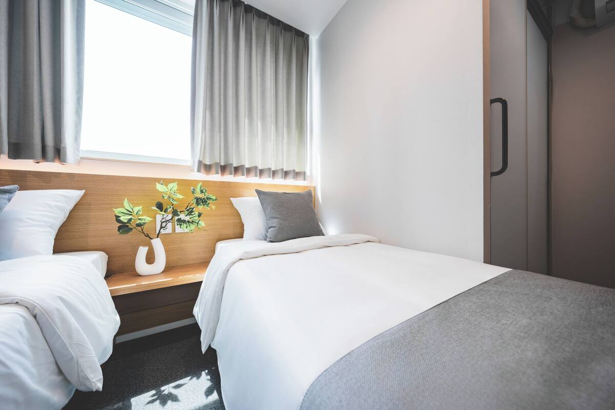 Habitación de hotel con dos camas, cabecera de madera, ventana con cortinas, mesita de noche con un jarrón de flores y un armario.