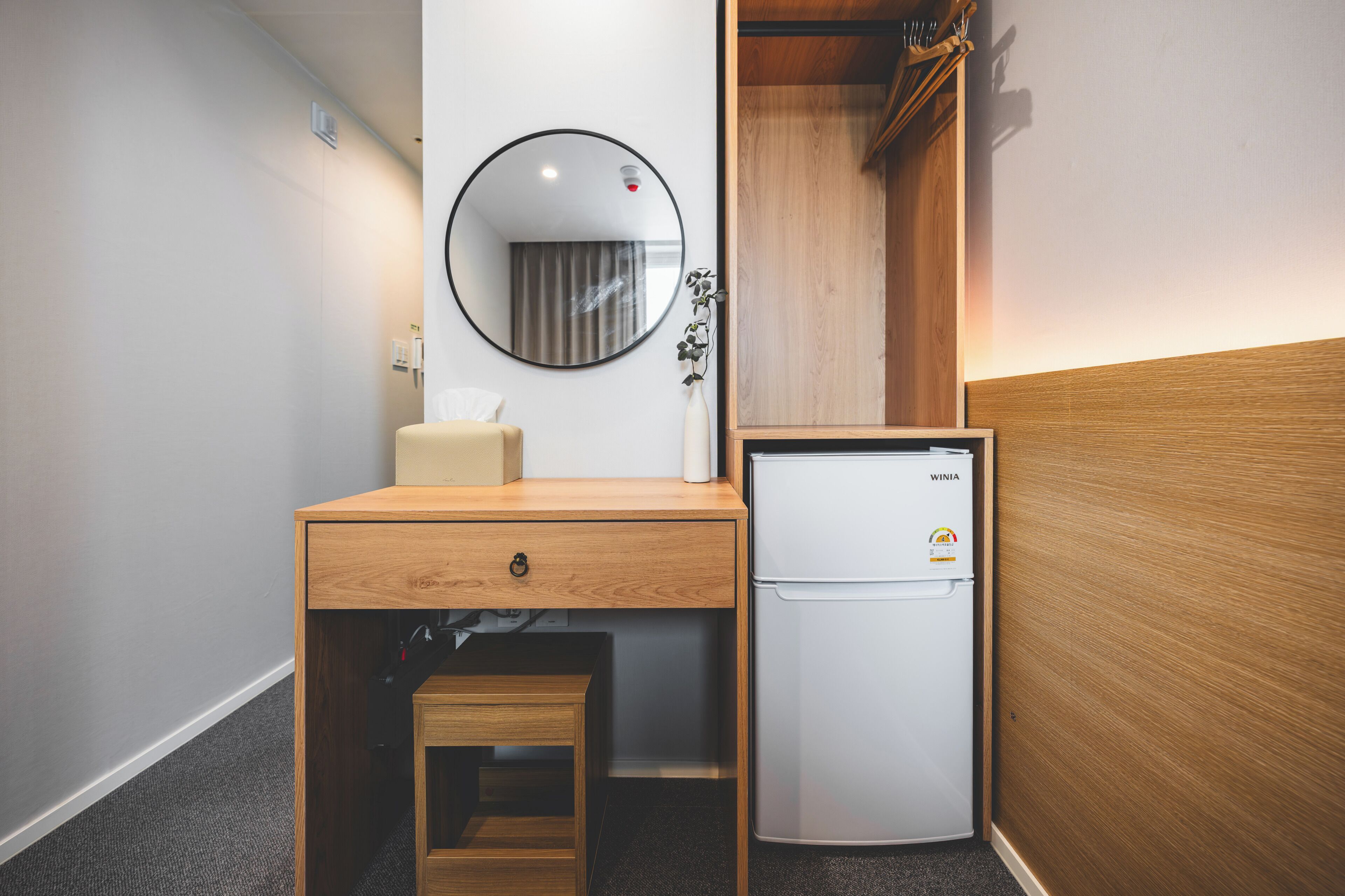 A compact room with a wooden desk, a small refrigerator, a mirror, and a bedside table.