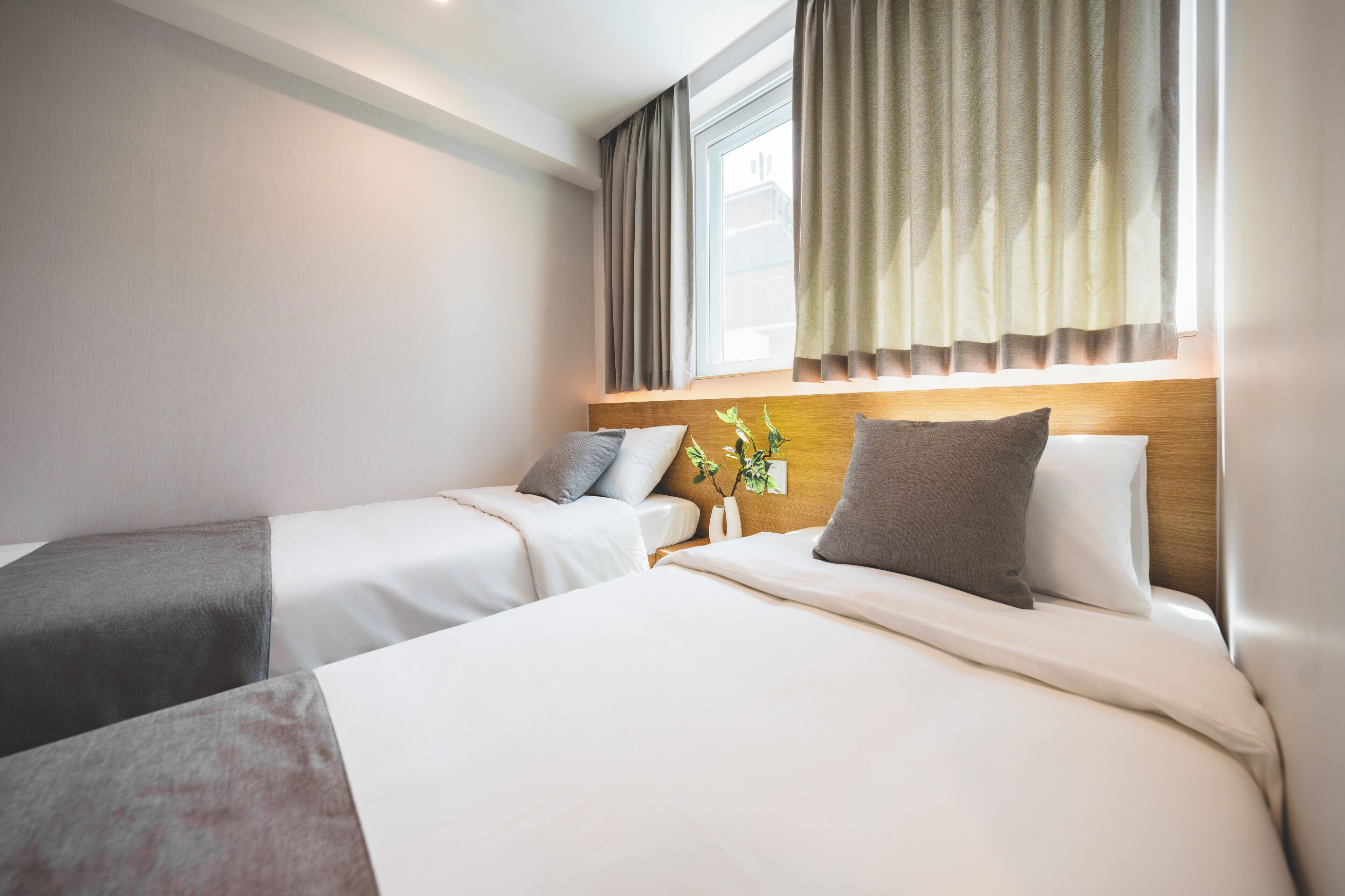 A hotel room with two beds, a window with curtains, a bedside table with a vase of flowers, and a wooden headboard.
