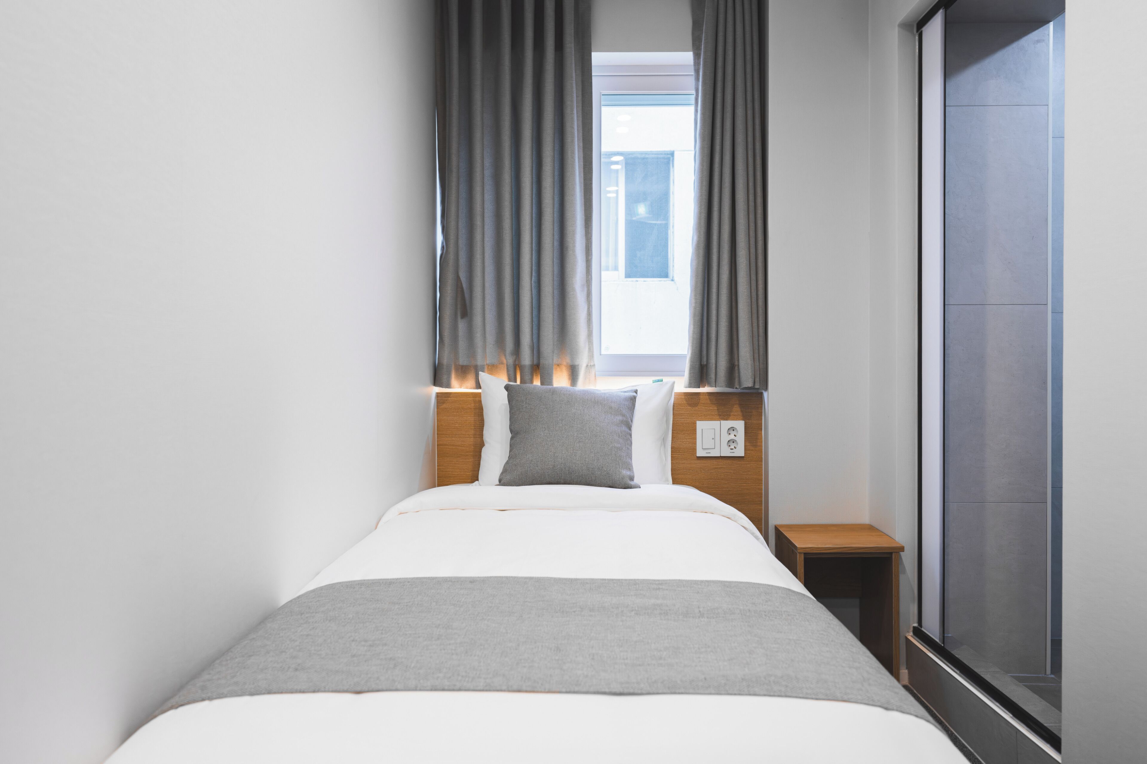A hotel room with a bed, a nightstand, a window with curtains, and a view of the outside.