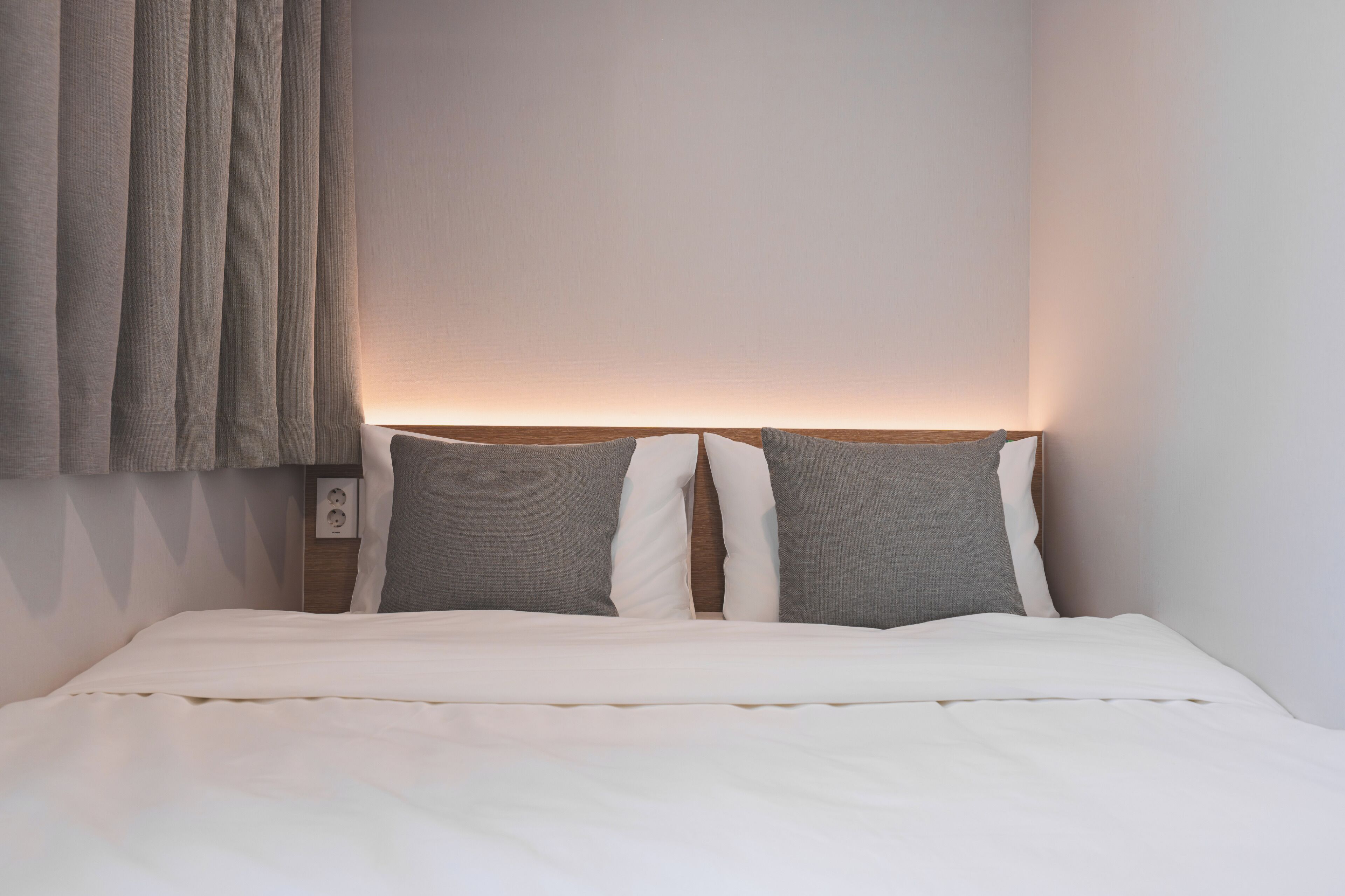 Una cama bien hecha con ropa blanca y dos almohadas, cabecera de madera y lámpara fijada a la pared.