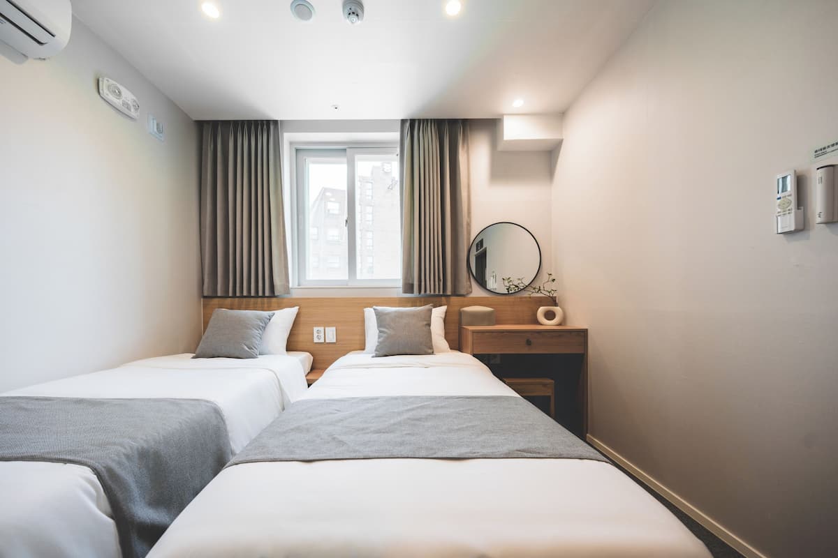 Habitación de hotel con dos camas, una ventana con cortinas, una mesita de noche con lámpara y un aire acondicionado instalado en la pared.