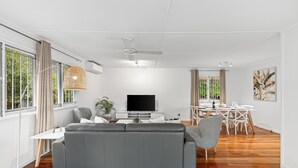 Una sala moderna con un sofá gris, un televisor de pantalla plana, una zona de comedor con mobiliario blanco y ventanas grandes con cortinas.