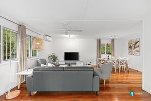 A modern living room with a grey sofa, a flat-screen TV, a dining area with white furniture, and large windows with curtains. - Stylish Mid Century Pet Friendly Home Near City (Tarragindi)