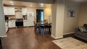 Una cocina moderna con armarios blancos, un comedor con mesa y sillas de madera, y un sofá gris en la sala.