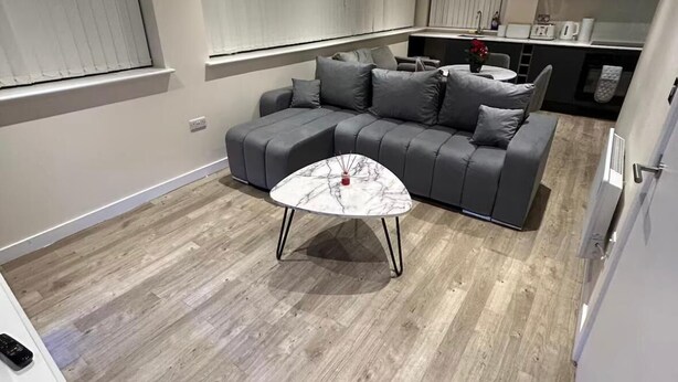 A modern living room with a grey sectional sofa, a white marble coffee table, and a kitchen area in the background. - Modern 1-Bed Penthouse — Steps from Old Trafford (Stretford)