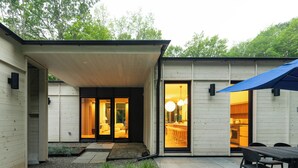 A modern house with a covered patio, a dining area with a table and chairs, and a kitchen visible through the open door. - New! The Woods ;  Luxury Home, Pool,  Hot Tub, Fire pit (Saugerties)
