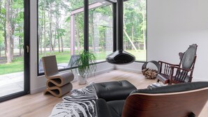 A modern living room with large windows, a fireplace, and a view of a forest. - New! The Woods ;  Luxury Home, Pool,  Hot Tub, Fire pit (Saugerties)