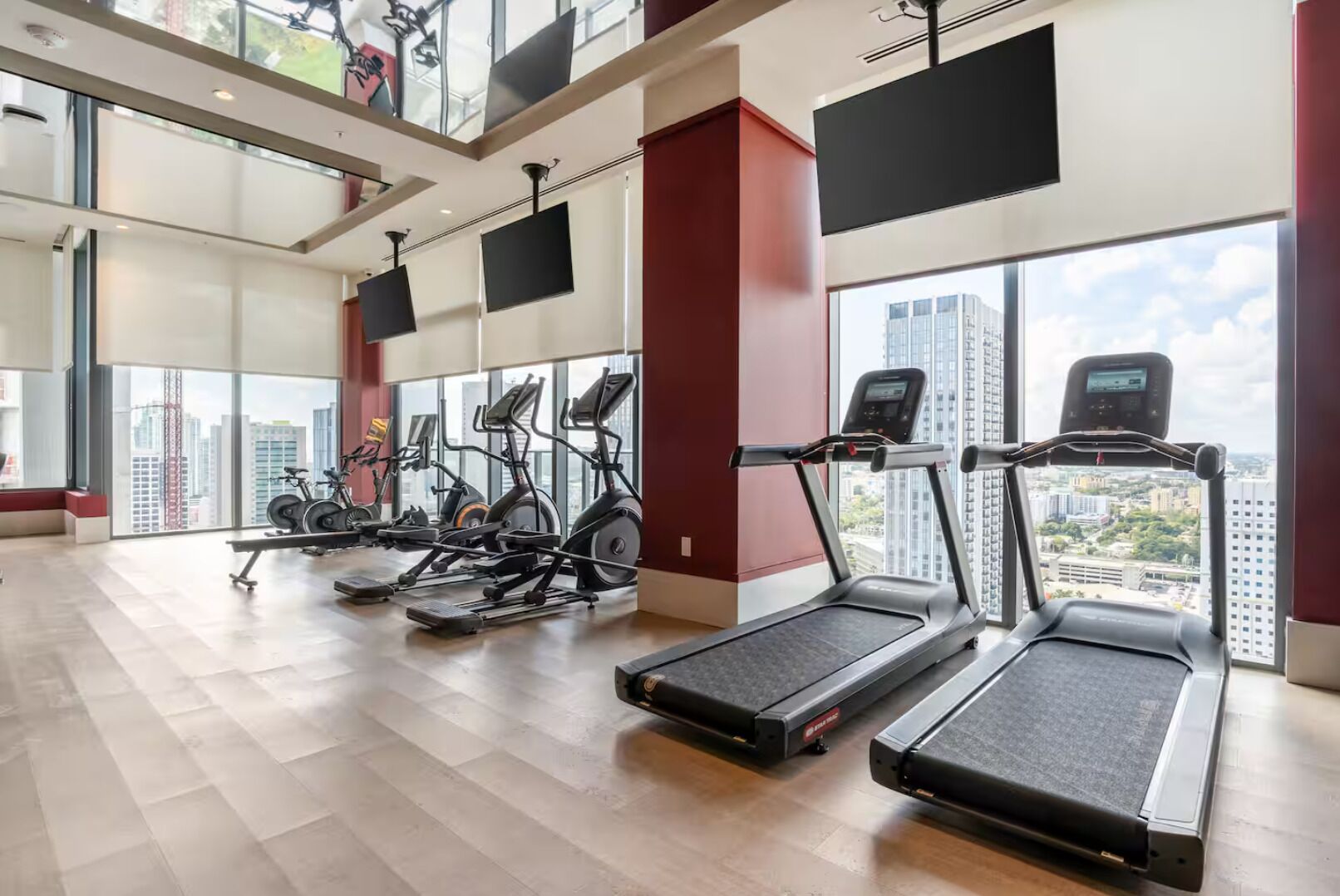A modern gym with multiple cardio machines, large windows offering city views, and a spacious layout.