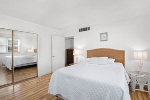 A bedroom with a large bed, a wooden headboard, a bedside table with a lamp, a mirror, and a wardrobe. - 🌅 Private Rooftop Deck Retreat | 2BR Coastal Condo at Gulf Winds Resort (Saint Pete Beach)