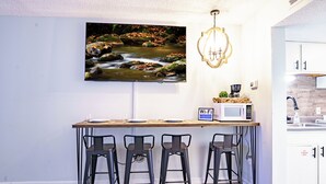 Una cocina con una barra de madera, tres taburetes de bar, un microondas y un cuadro grande con una imagen de un arroyo.