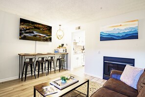 A modern living room with a wooden coffee table, a brown leather sofa, a fireplace, and a dining area with a kitchen in the background. - Gatlinburg Mountain Charm, Walk to DTWN, Sleeps 4, Elevator on Premises (Gatlinburg)