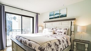 Un dormitorio con una cama grande, vistas a través de un ventanal corredero, una mesita de noche pequeña con una lámpara y un cuadro sobre la cama.