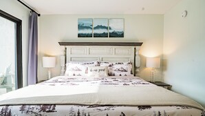 Una cama bien hecha con un edredón con motivos de montaña, dos lámparas de noche y un cabecero con un panel decorativo.