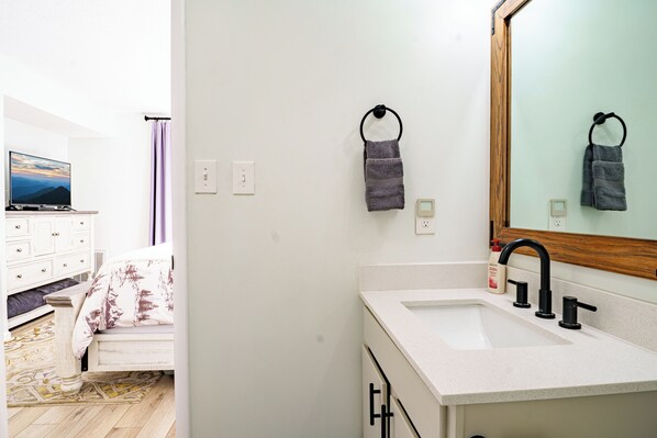 A modern bathroom with a white sink, black faucet, and a wooden-framed mirror. There are two towels hanging on hooks and a television in the adjacent room. - Gatlinburg Mountain Charm, Walk to DTWN, Sleeps 4, Elevator on Premises (Gatlinburg)