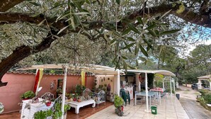 Een terras buiten met een overdekte zithoek, planten in potten en een witte tafel.