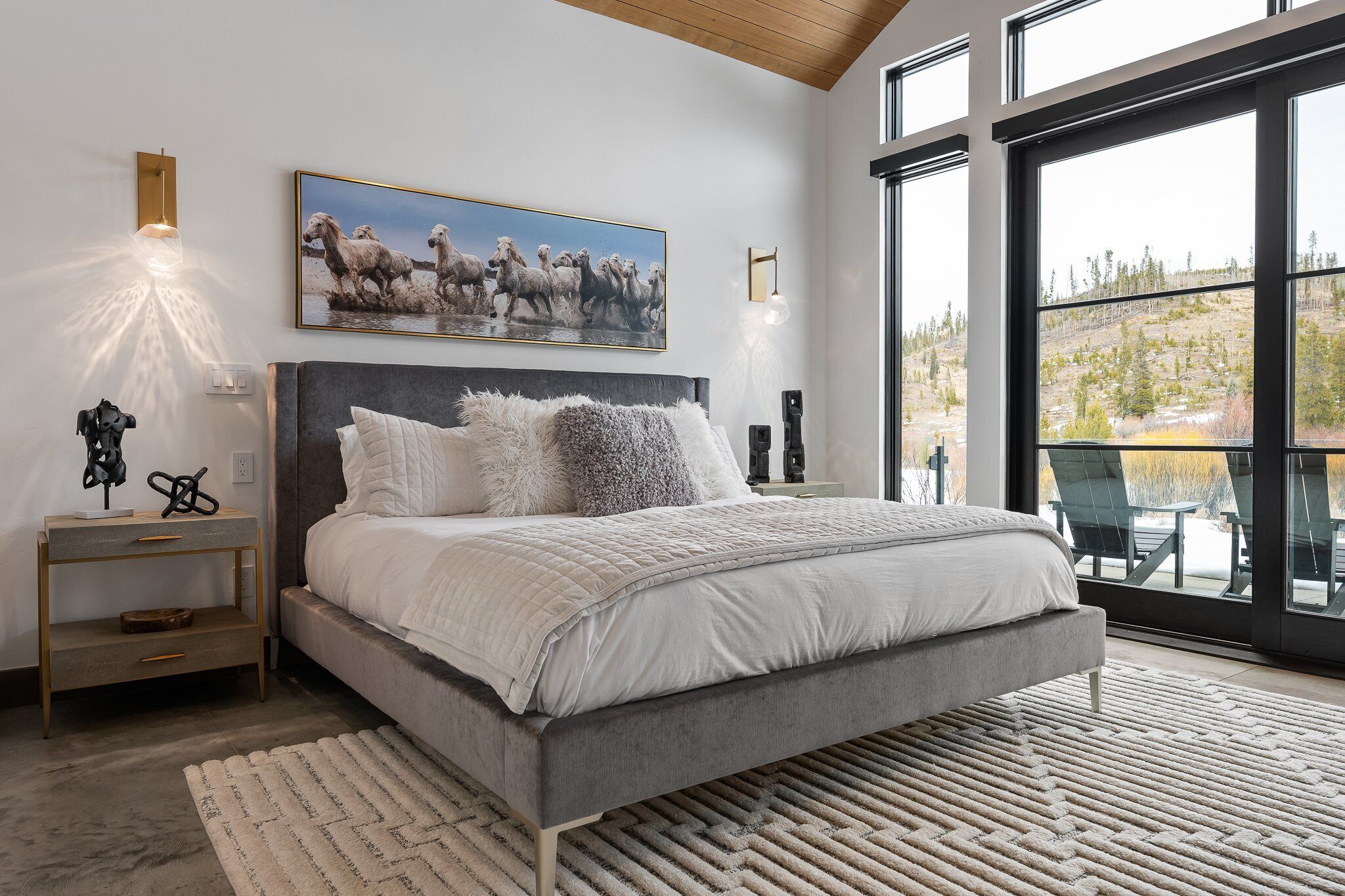 A modern bedroom with a large bed, a nightstand, a wall-mounted light, and a large window with a view of trees.