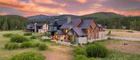 A large, modern house with multiple sections, surrounded by a natural landscape and mountains.