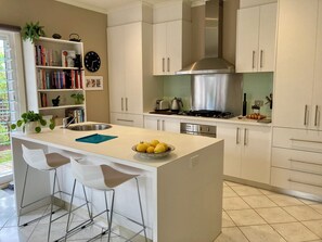 A modern kitchen with white cabinetry, a central island with bar stools, a stainless steel stove, and a bookshelf filled with books and decorative items.